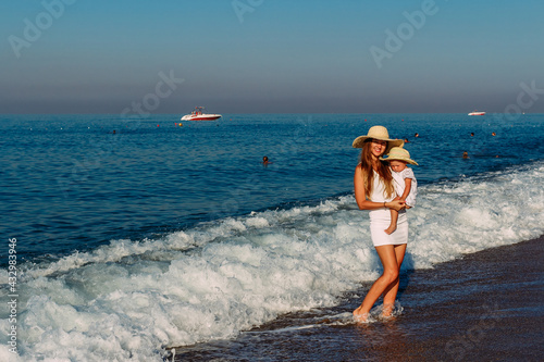 Smiling woman in hat holds in her arms a one-year-old baby girl standing on the sea in the waves in the summer