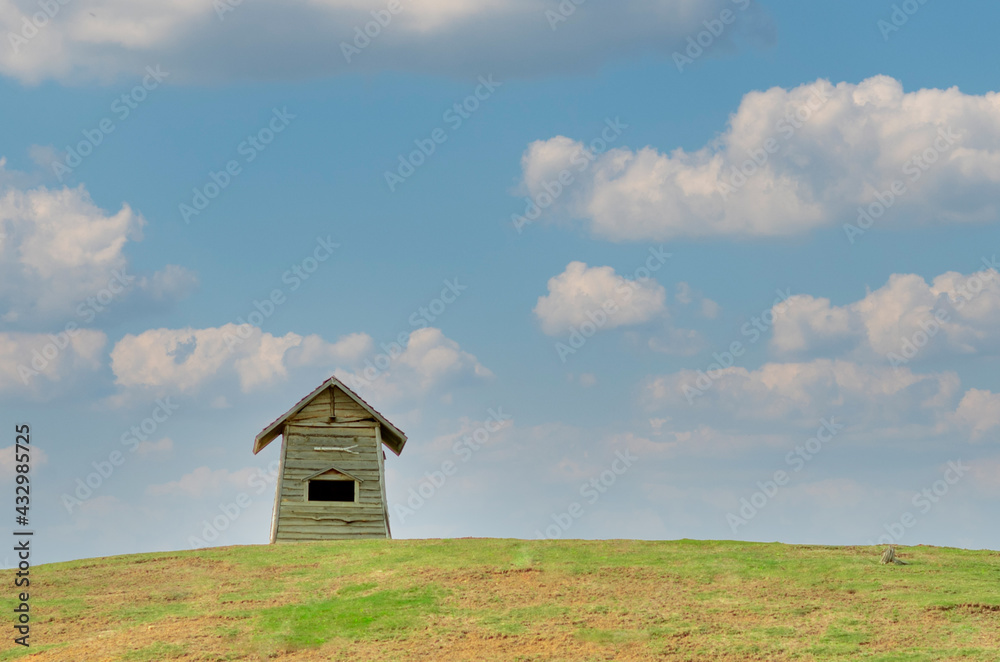 A small wood warehouse storage barn house building on top of hill and green turf grass meadow field with blue sky and white cloud as background in pastel color tone. Spring season. A small wood warehouse storage barn house building on top of hill and green turf grass meadow field with blue sky and white cloud as background in pastel color tone. Spring season.
