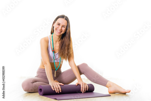 Young sporty female yoga instructor in bright white yoga studio, smiling cheerfully while rolling the yoga mat after yoga session.