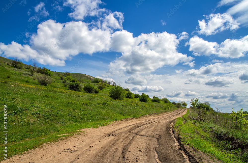 Dirt road at the foot of the mountain. A hillside with young greenery against the background of a sky with cumulus clouds. 