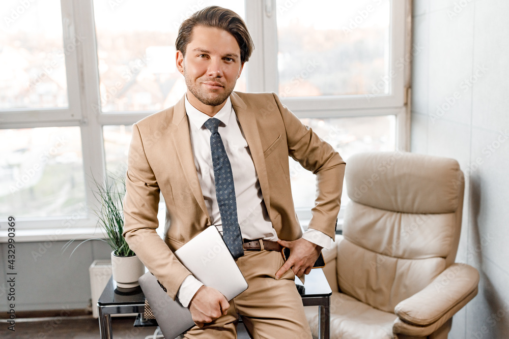 Successful male businessman or lawyer standing in his office with french window, near desk with laptop, looking at the camera and smiling. Confident male wearing stylish suit and tie Successful male businessman or lawyer standing in his office with french window, near desk with laptop, looking at the camera and smiling. Confident male wearing stylish suit and tie