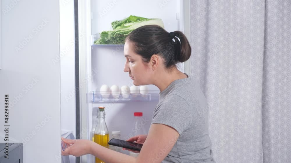 Concentrated young housewife in grey t-shirt makes list of food products to buy looking inside open refrigerator in kitchen side view Concentrated young housewife in grey t-shirt makes list of food products to buy looking inside open refrigerator in kitchen side view