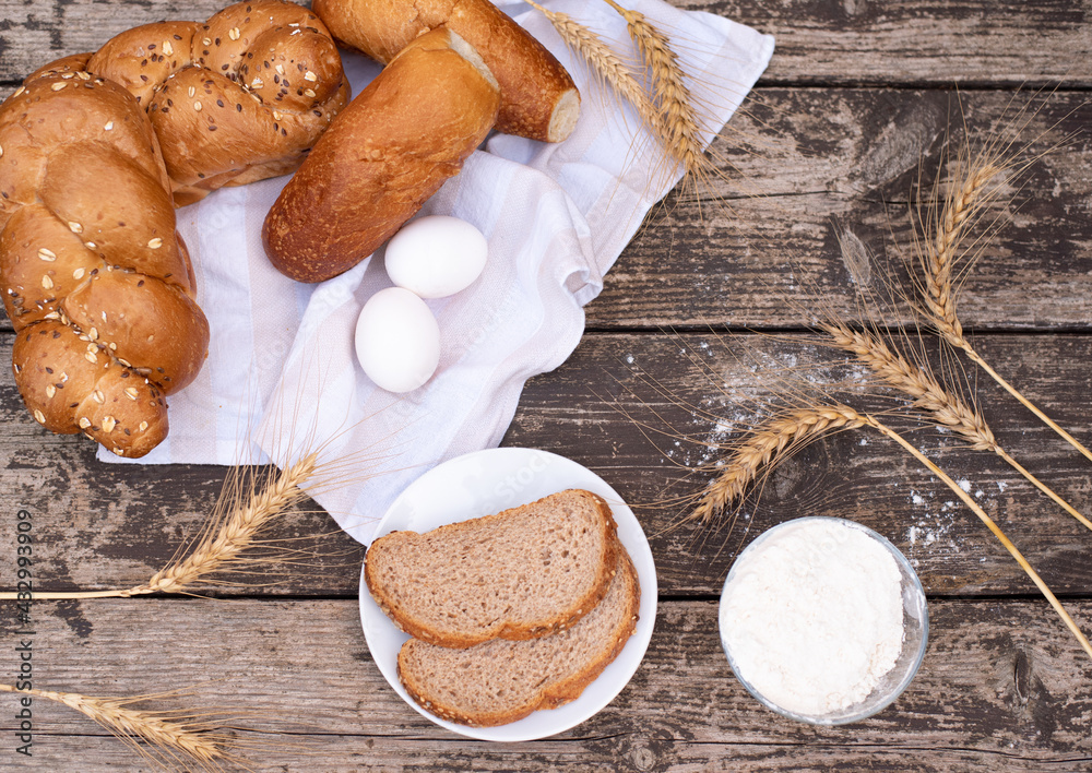 Fototapeta premium Fresh crispy bread and two eggs on white napkin with spikelets of wheat, bowl of flour, bread on plate on wooden table. Harvest season holiday World Bread Day. Bread is the staff of life. Top view.