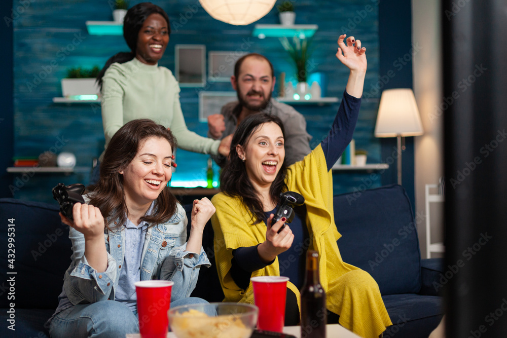 Fototapeta premium Cheerful women celebrating victory while playing video games with friends using wireless controller. Group of mixed race friends playing games while sitting on sofa in living room late at night.