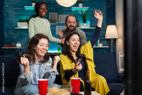Cheerful women celebrating victory while playing video games with friends using wireless controller. Group of mixed race friends playing games while sitting on sofa in living room late at night.