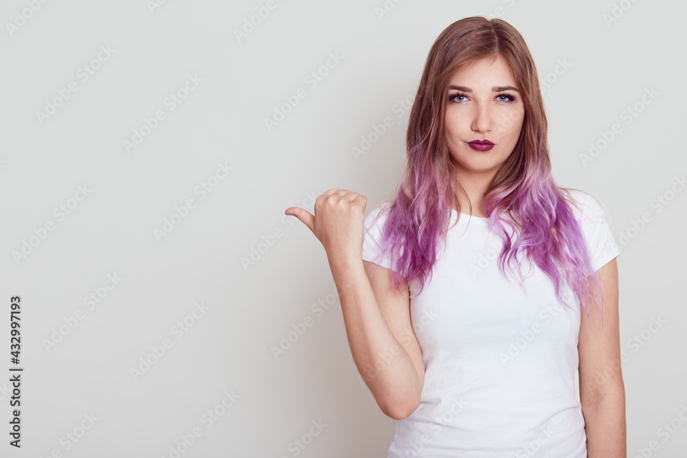 Serious woman wearing casual white t shirt looking at camera with strict expression, pointing thumb aside, showing free space for advertisement, posing isolated over gray background. Serious woman wearing casual white t shirt looking at camera with strict expression, pointing thumb aside, showing free space for advertisement, posing isolated over gray background.