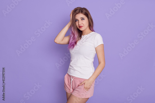 Self confident beautiful female wearing white t shirt and pink shorts standing, keeping hand on head, looking at camera, posing isolated over purple background. © sementsova321