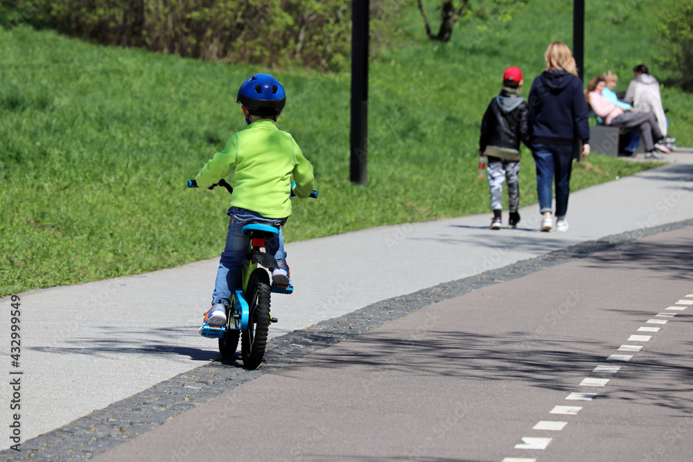Obraz premium Little boy rides a bicycle on a street in summer park on walking people background