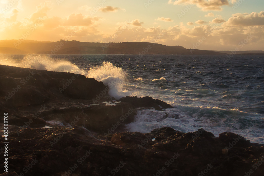 Waves crashing over rocks lit up with golden light from the setting sun on a beach in Qawra, Malta.