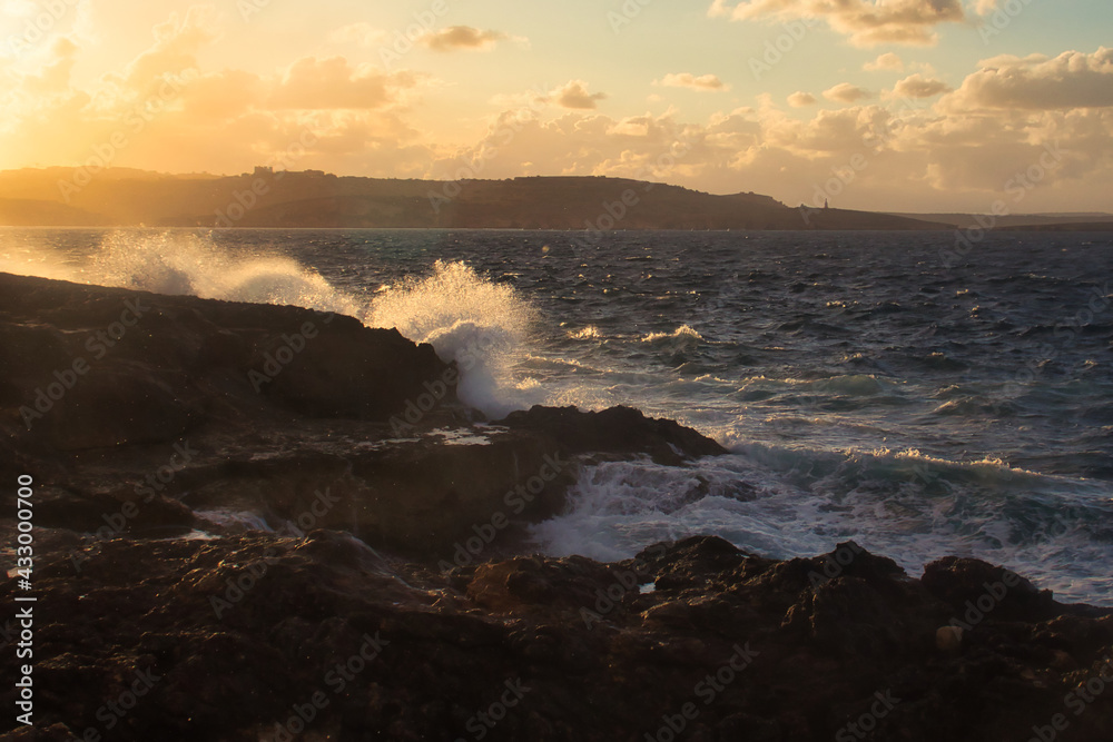 Obraz premium Waves crashing over rocks lit up with golden light from the setting sun on a beach in Qawra, Malta.