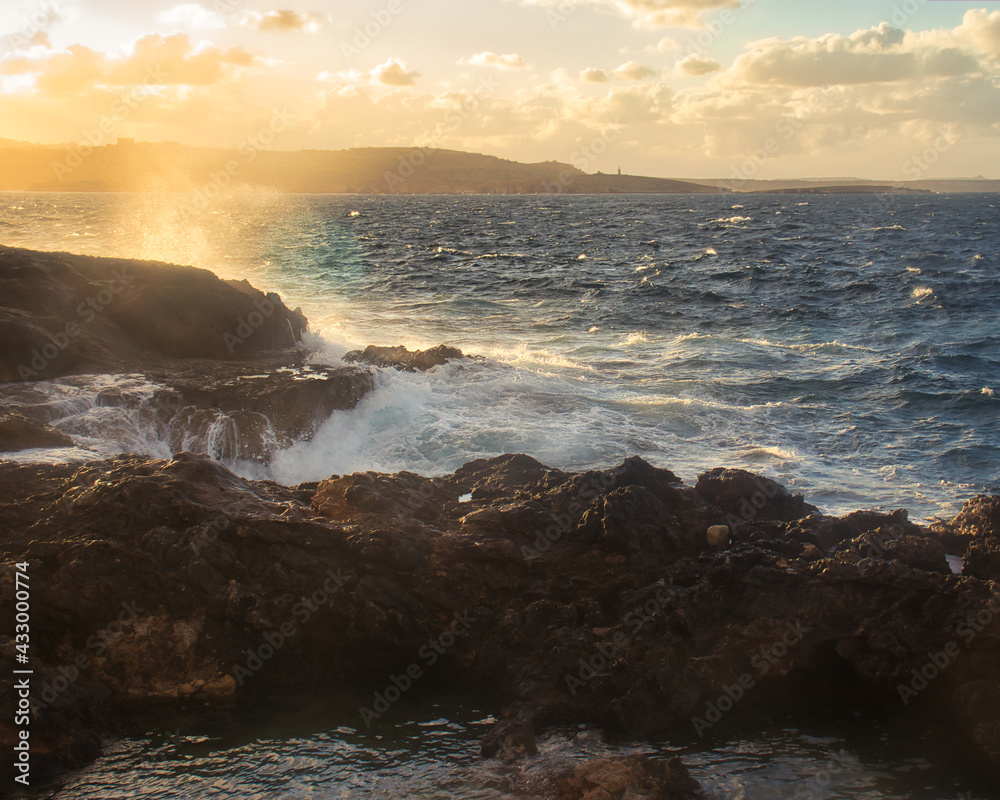 Obraz premium Golden light hitting water spraying over rocks on a beach in Qawra, Malta.