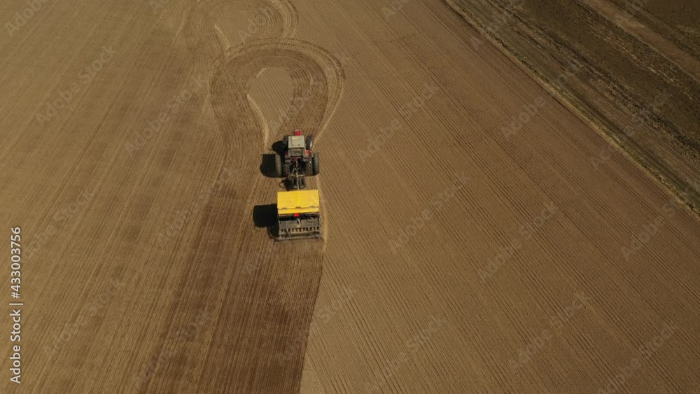 Tracking aerial view of a red double wheeled tractor sowing seeds on a agricultural field with a yellow seeder during spring time. Tractor is turning around at the end to start a new row. Tracking aerial view of a red double wheeled tractor sowing seeds on a agricultural field with a yellow seeder during spring time. Tractor is turning around at the end to start a new row.