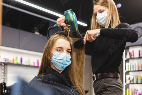 Master woman hairdresser in a medical mask dries the girl's hair with a hairdryer and combs after washing in a beauty salon. Covid-19 pandemic and remedies