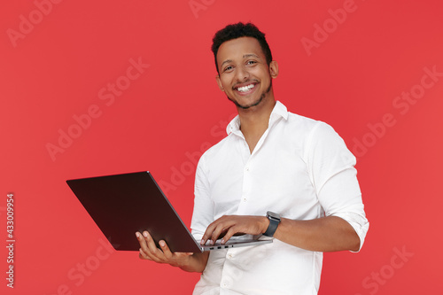 Smiling african american young man holding laptop over red background.