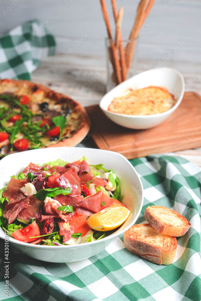 Prosciutto and arugula salad in a white bowl and cherry tomatoes pizza over wooden background. Red plaid tablecloth. Prosciutto and arugula salad in a white bowl and cherry tomatoes pizza over wooden background. Red plaid tablecloth.