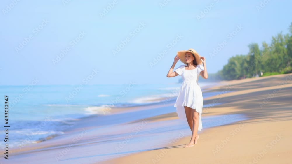 Happy woman in white summer dress and straw hat walking barefoot along the beach on sea tides background, sunny day in tropical island beach Happy woman in white summer dress and straw hat walking barefoot along the beach on sea tides background, sunny day in tropical island beach