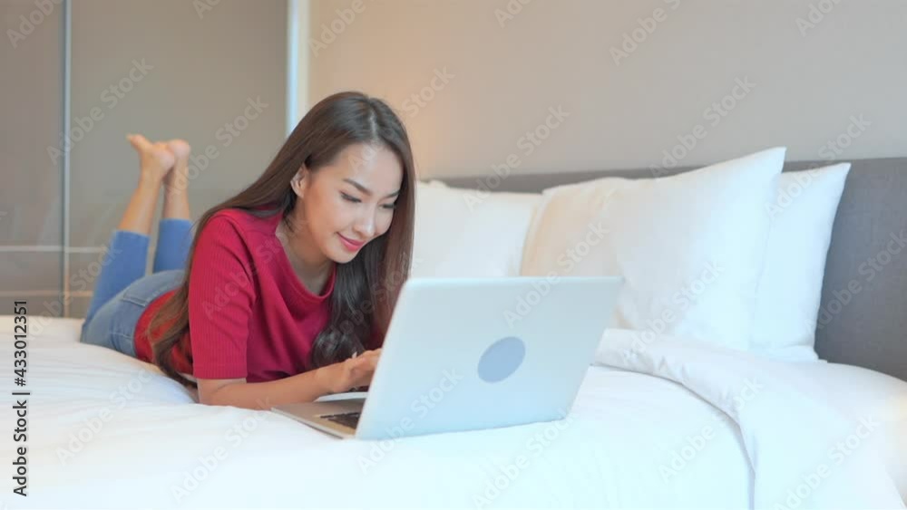 Using notebook on vacation. Slow-motion of relaxed Asian woman using her laptop computer lying on the bed with her legs raised at hotel room wearing casual clothes Using notebook on vacation. Slow-motion of relaxed Asian woman using her laptop computer lying on the bed with her legs raised at hotel room wearing casual clothes