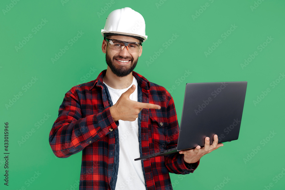 Smiling bearded engineer or constructor man in casual outfit pointing finger on computer over green background. - obrazy, fototapety, plakaty 