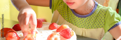 Fotografie cute little caucasian seven year old kid girl at home at the table decorates a homemade baking cake with strawberries on her birthday on a sunny day