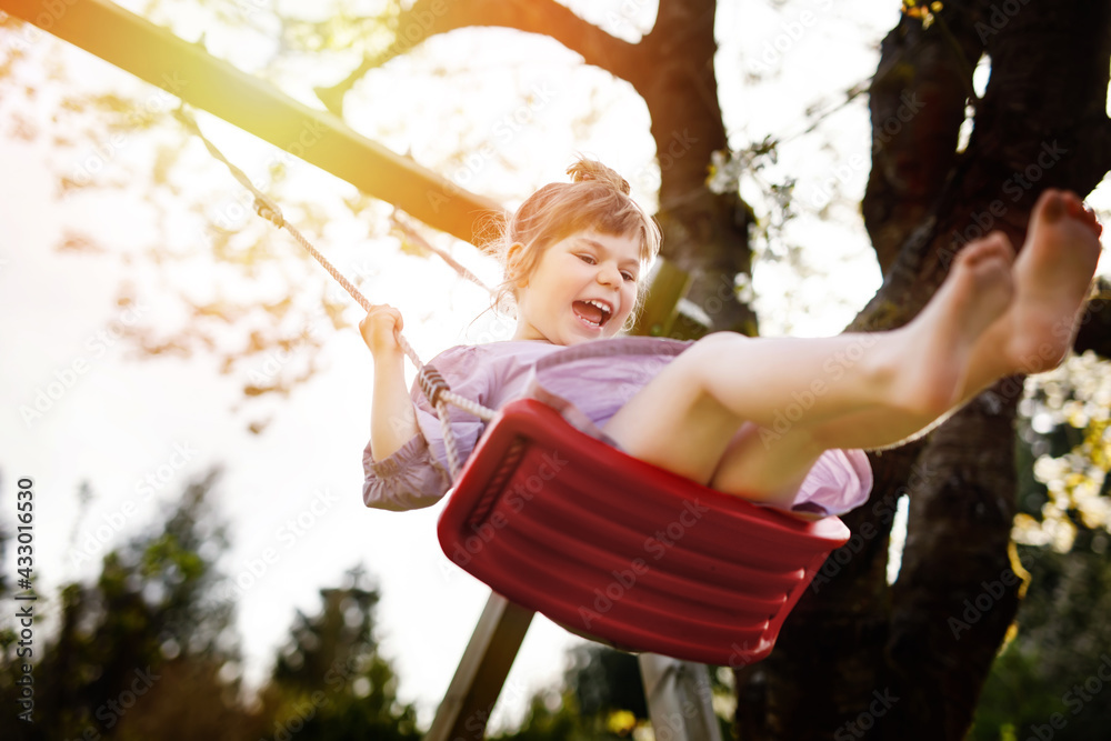 Happy little toddler girl having fun on swing in domestic garden. Smiling positive healthy child swingingon sunny day. Preschool girl laughing and crying. Active leisure and activity outdoors.
