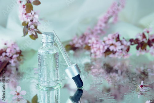 Closeup of wet glass bottle, glass-dropper and blurred tender pink flowers on the background with wet reflecting surface underneath. Herbal medicine and cosmetics concept.