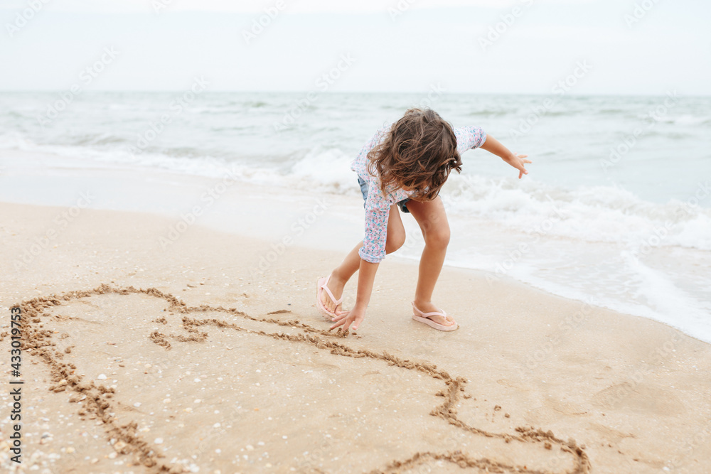 Five years cute curly caucasian girl painting on sand on the beach.
