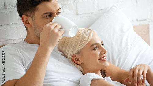 tattooed man drinking coffee while lying with pleased blonde woman in bed