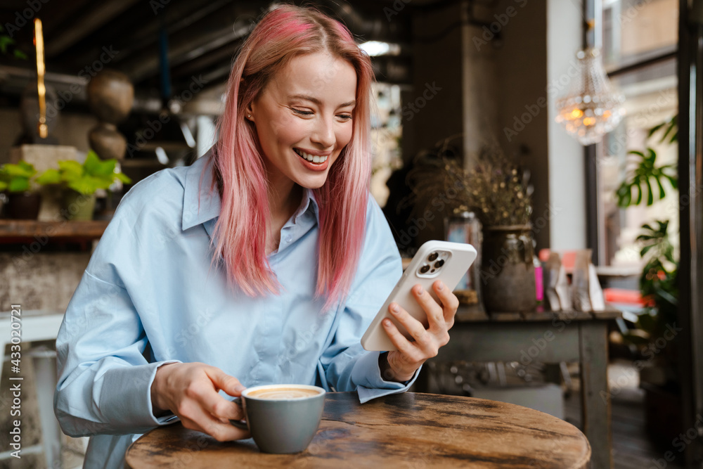 Young smiling woman using mobile phone while drinking coffee in cafe