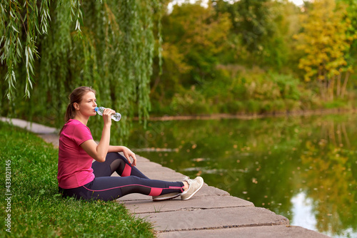 young woman in sportswear sits on the river bank in the park and drinks water © Hanna