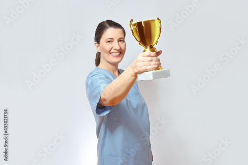 Middle aged nurse in a light blue scrub smiling proudly and holding a golden trophy up, white background, copy space. photo