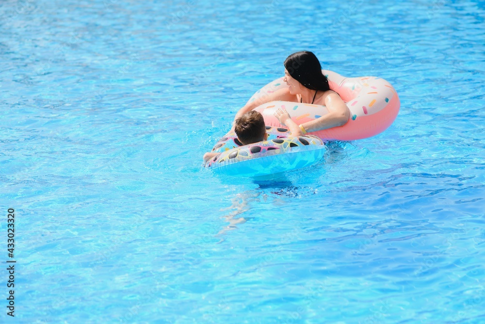 Happy family, young active mother and adorable curly little baby having fun in a swimming pool, child learning to swim in an inflatable toy ring, enjoying summer vacation at a tropical resort