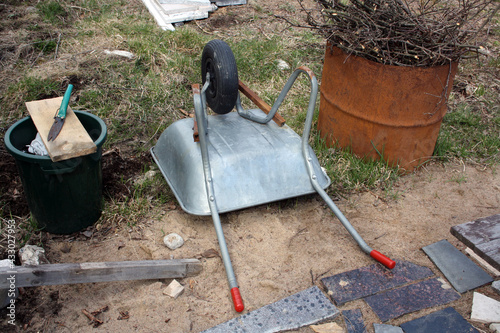 Construction wheelbarrow turned upside down and a rusty barrel with branches.