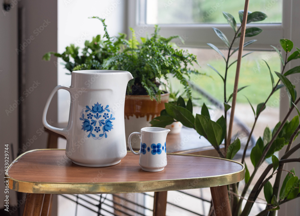 Mid-century modern water-jug and small pitcher on a wooden table before the window with plants Mid-century modern water-jug and small pitcher on a wooden table before the window with plants