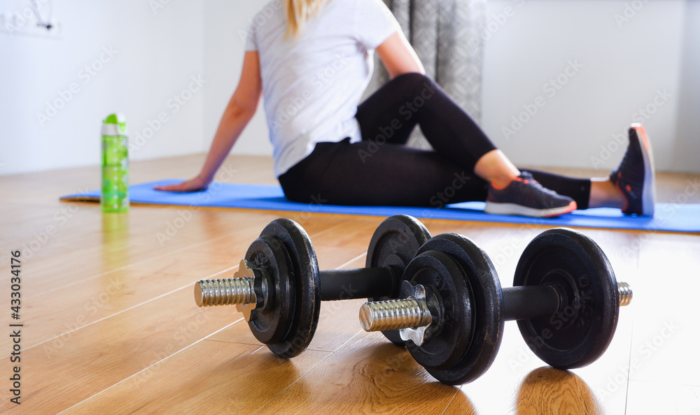 Home workout. Unrecognizable blonde woman in black leggings and sneakers doing stretching after strength training with dumbbells Woman on a pink mat at home. Focus on the dumbbells in the foreground. Home workout. Unrecognizable blonde woman in black leggings and sneakers doing stretching after strength training with dumbbells Woman on a pink mat at home. Focus on the dumbbells in the foreground.