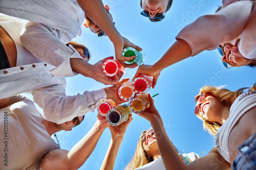 Photography Low angle view of friends having fun at poolside summer party, clinking glasses with colorful summer cocktails near hotel swimming pool