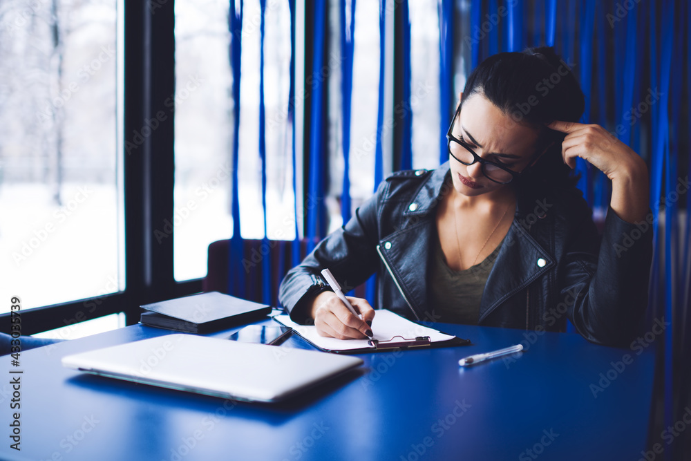 Pensive entrepreneur sitting at table and taking notes in document in clipboard