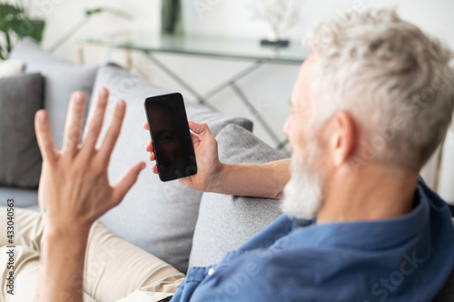 Grey haired senior middle-aged man using smartphone for video call, phone with black empty screen in his hand, back view, mature man waving hello to blank screen, video call, virtual meeting. Mockup