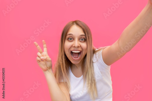 A young pretty caucasian impressed excited smiling cheerful blonde woman in a white t-shirt takes selfie showing a peace gesture with her v-shaped hand isolated on a bright color yellow background