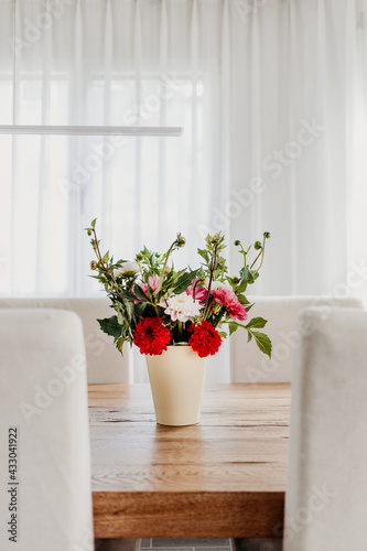 Dahlia flowers bouquet in a vase on wooden dining table. Modern room interior, bright and airy. © nruedisueli