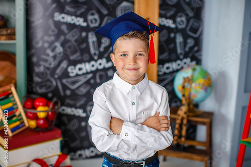 Portrait of a little schoolboy in a white shirt and a graduate cap posing in a school classroom