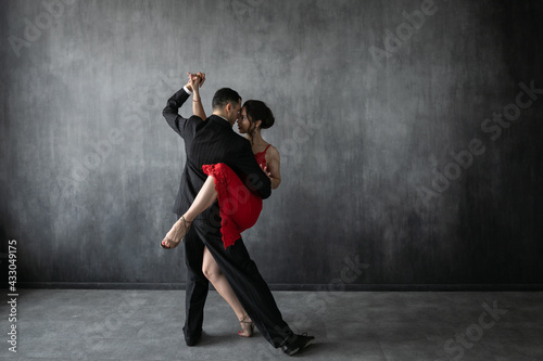 Couple of professional tango dancers in elegant suit and dress pose in a dancing movement on dark background. Attractive man and woman dance looking eye to eye.