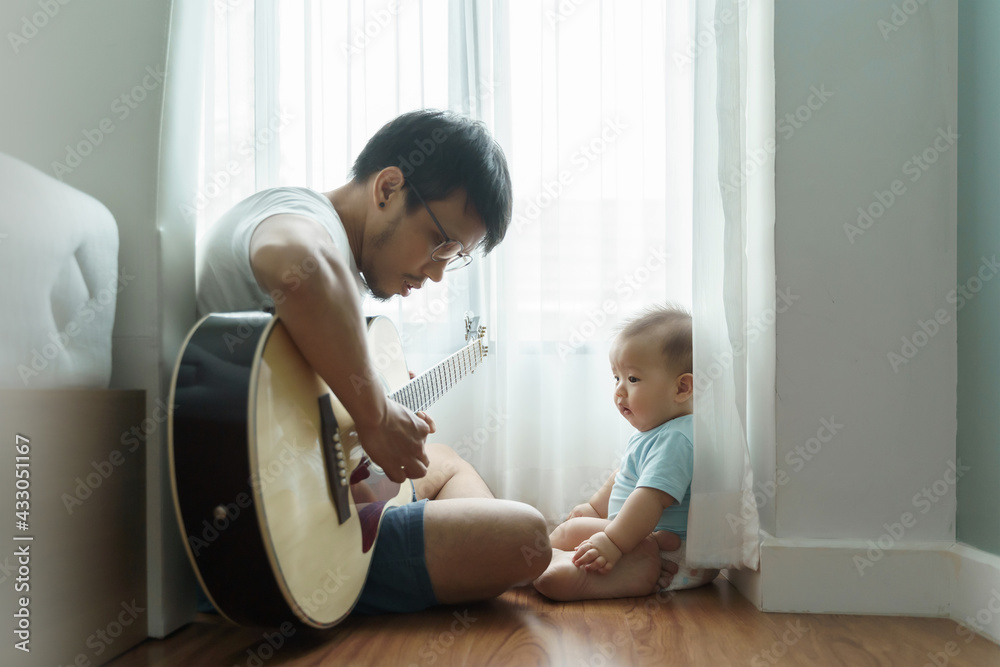 Asian Dad sitting on wood floor playing acoustic guitar with his little Cute son together during quarantine. Asian Dad sitting on wood floor playing acoustic guitar with his little Cute son together during quarantine.