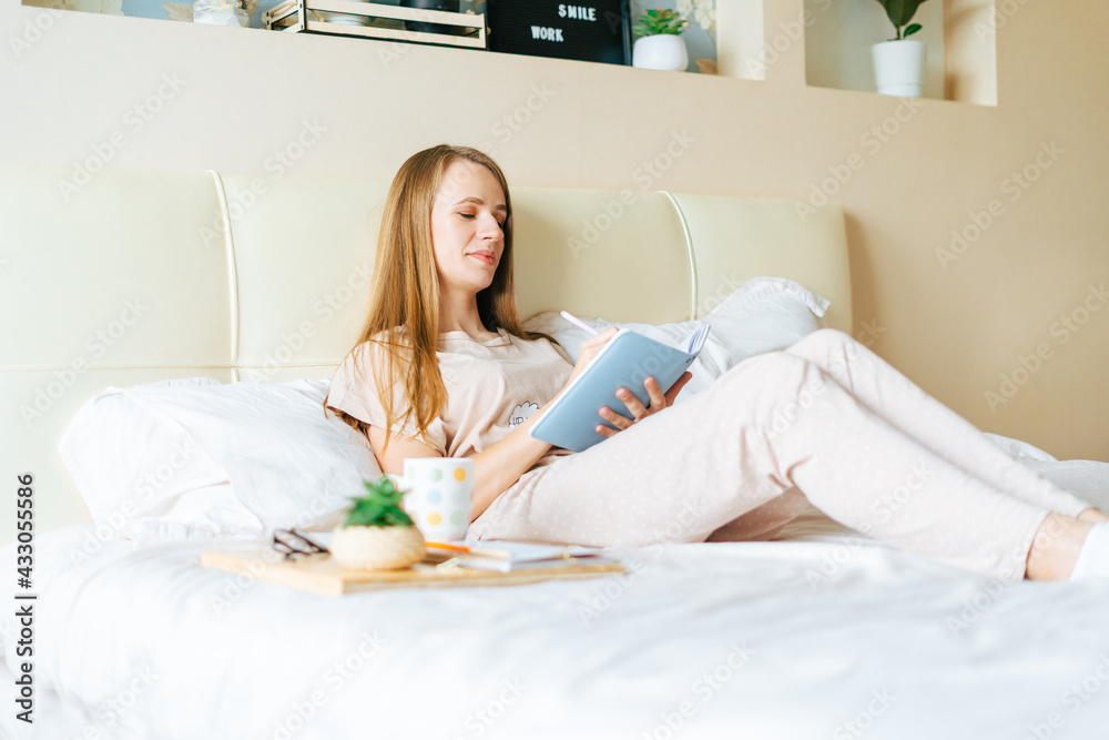 Close up of relaxed cheerful woman reading and writing in notebook, working from home on bed and drinking coffee. Blonde girl Enjoying free leisure time with book. Distance learning and education. Close up of relaxed cheerful woman reading and writing in notebook, working from home on bed and drinking coffee. Blonde girl Enjoying free leisure time with book. Distance learning and education.