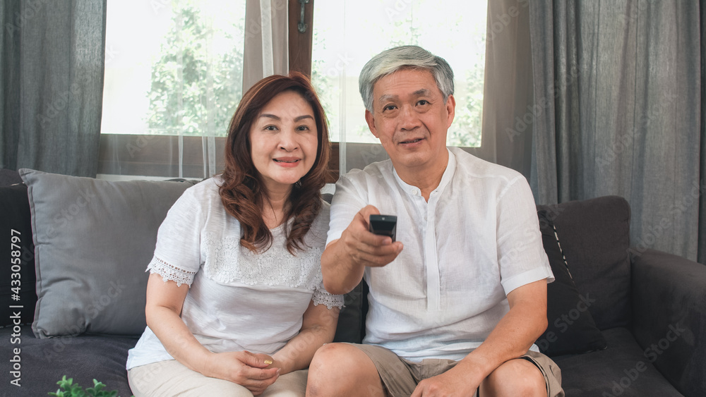 Obraz premium Asian elderly couple watching television in living room at home, sweet couple enjoy love moment while lying on the sofa when relaxed at home. Enjoying time lifestyle senior family at home concept.