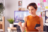 Female Architect In Office Working At Desk Making Online Purchase Using Credit Card On Mobile Phone