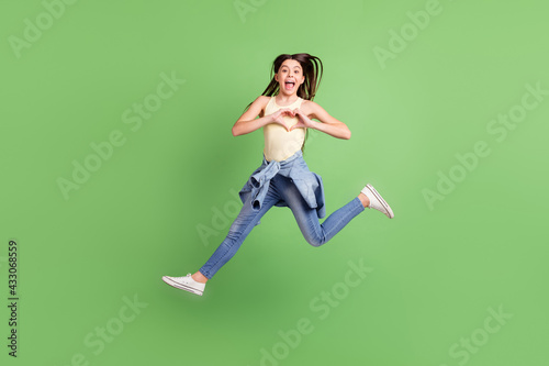 Fototapeta Naklejka Na Ścianę i Meble -  Full size photo of happy cheerful excited crazy little girl jumping showing heart sign isolated on green color background
