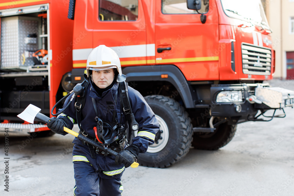 Portrait of serious and confident caucasian fireman standing and holding hammer, wearing special protective uniform in the truck background Portrait of serious and confident caucasian fireman standing and holding hammer, wearing special protective uniform in the truck background