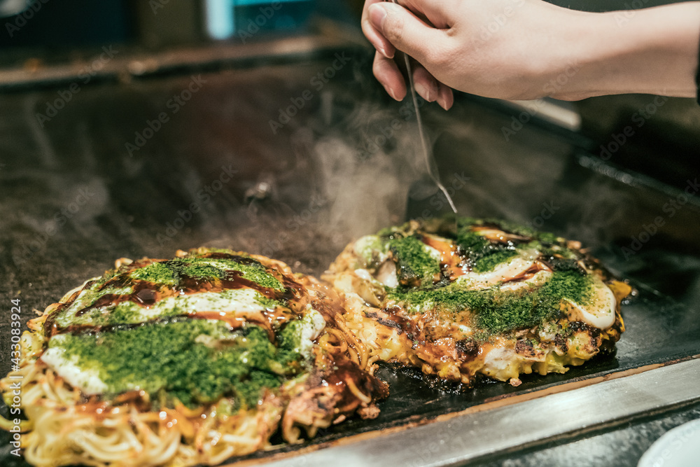 Naklejka premium Japanese style pancake okonomiyaki handmade on hot grill plate at restaurant. female hands customer ready to eat okonomiyaki on teppanyaki pan. local food in osaka japan.