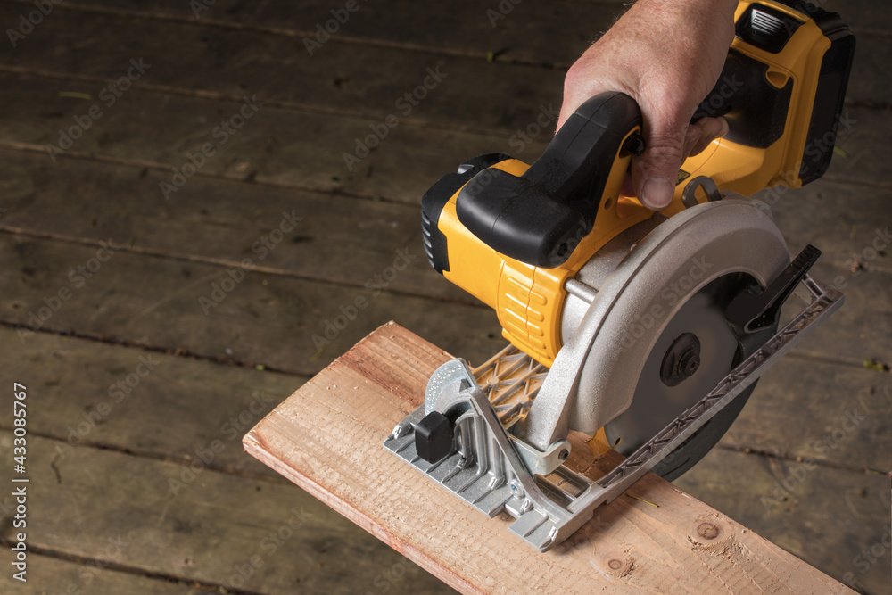 Male hand using an electric circular saw to cut a piece of wood on a dark construction site