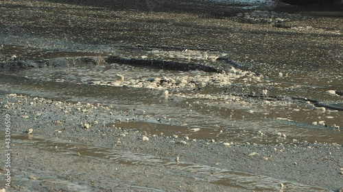 Close-up cars drive along a bad road with holes, muddy puddles and cracks. A crumbling road with potholes. Old asphalt pavement made in violation of the technology of work. Taxpayer money is wasted.